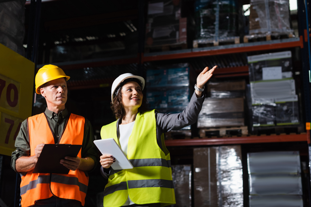 happy warehouse employee with tablet pointing away near supervisor in hard hat and safety vest - Photo, Image