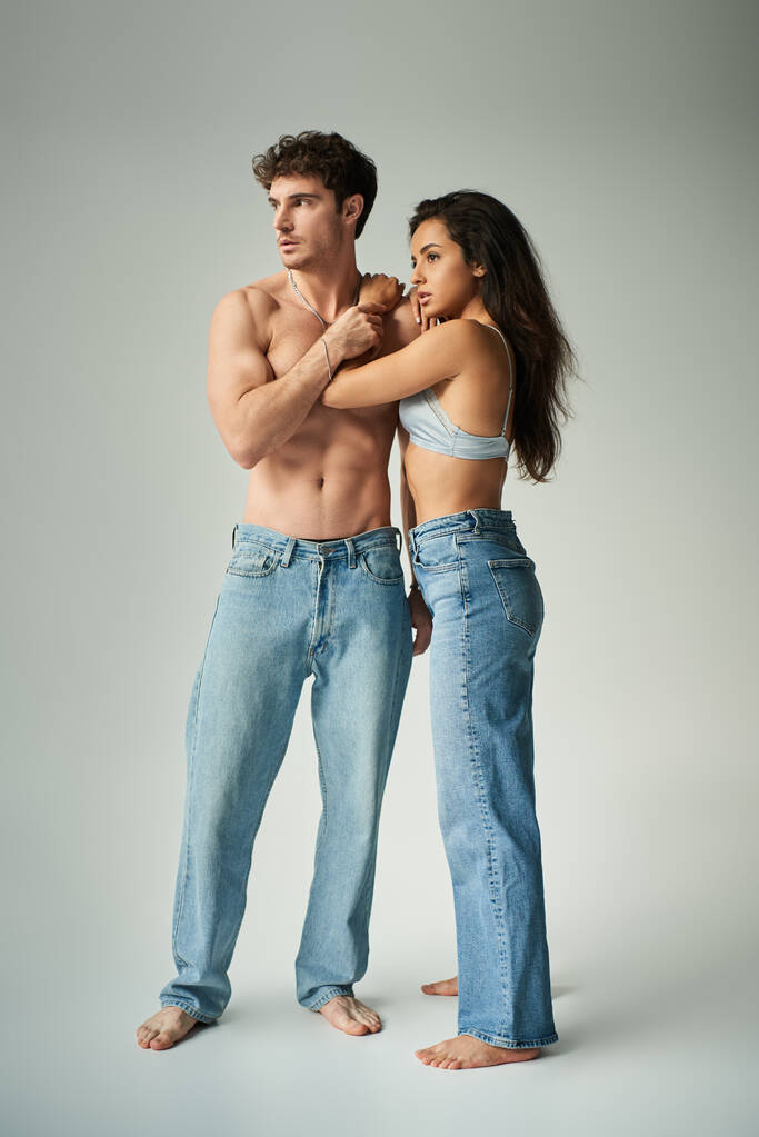 брюнетка молодая женщина в атласном бюстгальтере и джинсах обнимает мужчину без рубашки на сером фоне, привязанность - Фото, изображение
