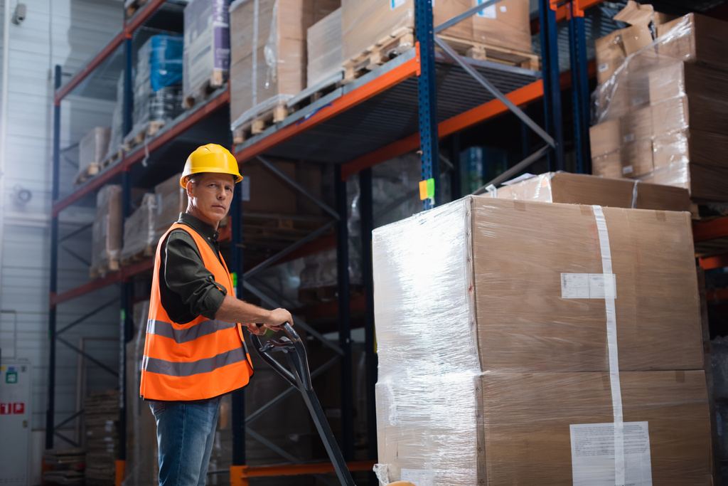 travailleur d'entrepôt d'âge moyen concentré en casque dur et gilet de sécurité transportant palette avec camion à main - Photo, image