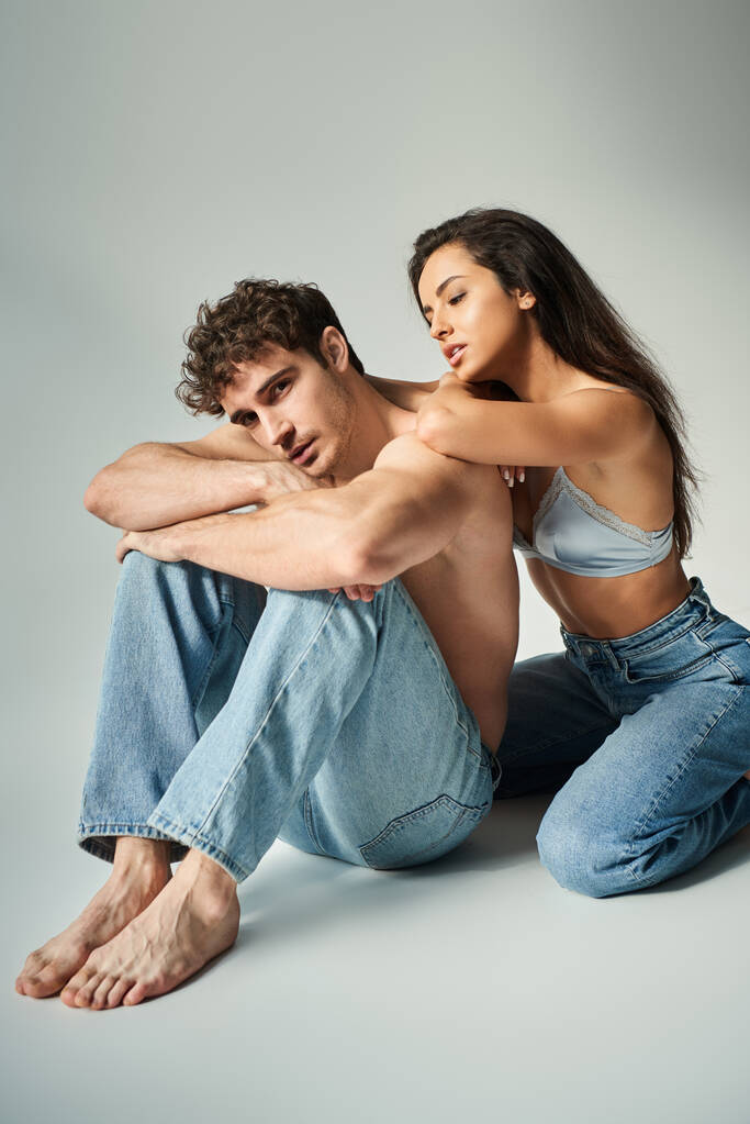 σέξι νεαρή γυναίκα σε σατέν σουτιέν και τζιν ποζάρουν με γυμνόστηθο άνδρα σε γκρι φόντο, εγγύτητα - Φωτογραφία, εικόνα