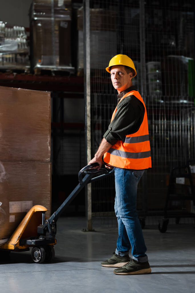 fuerte trabajador de almacén de mediana edad en chaleco de seguridad que transporta paleta pesada con camión de mano - Foto, imagen