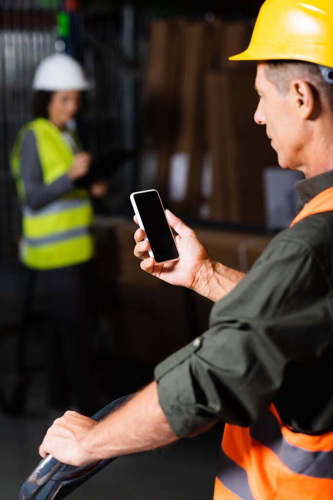 επόπτης στο γιλέκο ασφαλείας και κράνος χρησιμοποιώντας smartphone με εργαζόμενο στο παρασκήνιο της αποθήκης - Φωτογραφία, εικόνα