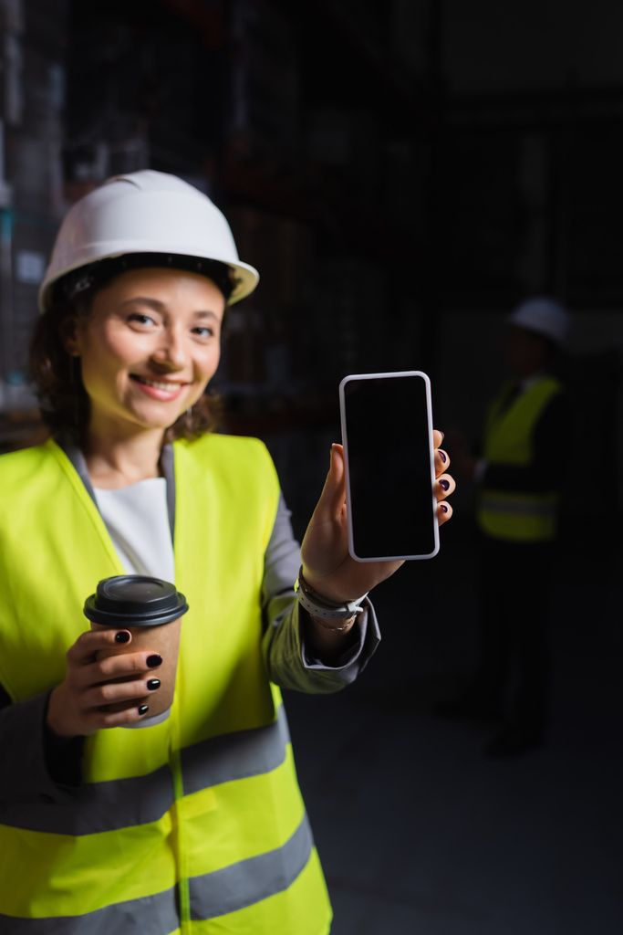smiling woman in hard hat holding smartphone and paper cup, happy warehouse employee on coffee break - Photo, Image
