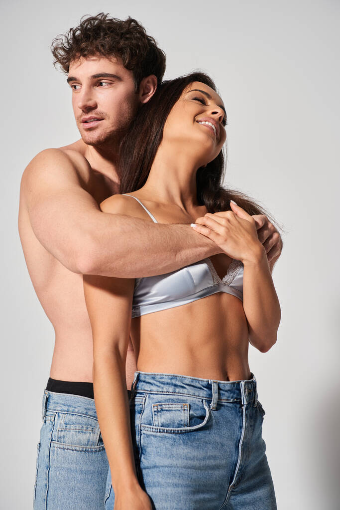 sexy en shirtloze man knuffelen verleidelijk jong en vrolijk vrouw op grijze achtergrond, saamhorigheid - Foto, afbeelding