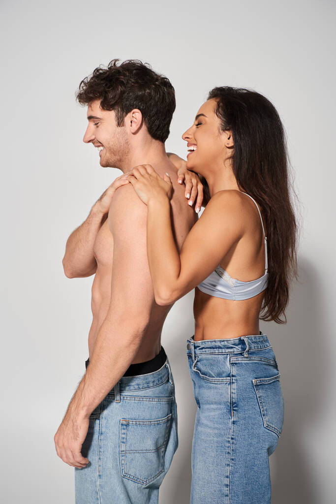 side view of happy young woman with brunette hair leaning on shirtless man on grey background - Photo, Image
