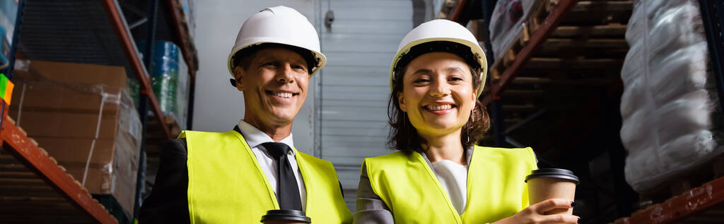 Cheerful logistics team with hard hats and coffee smiling during a break in warehouse, banner - Photo, Image
