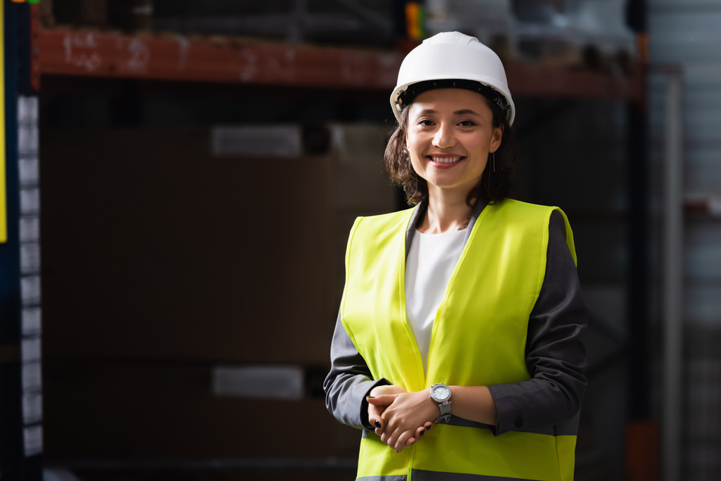 femme professionnelle gaie en gilet de sécurité et casque debout avec les mains dans les poches dans l'entrepôt - Photo, image
