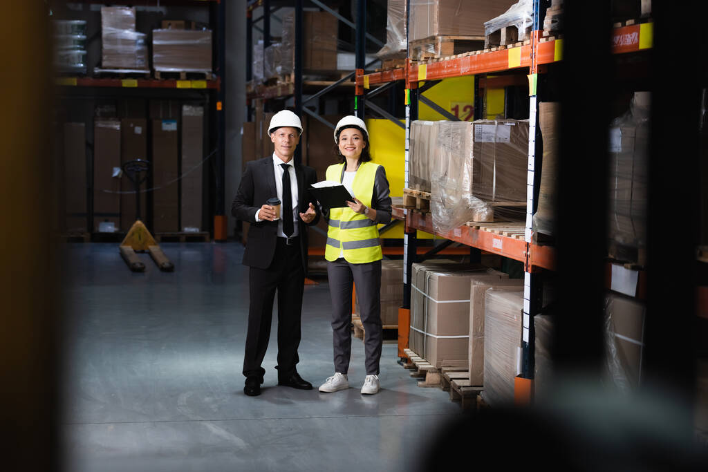 glimlachende zakenman in pak en harde hoed bespreken logistieke operaties met vrouwelijke werknemer - Foto, afbeelding