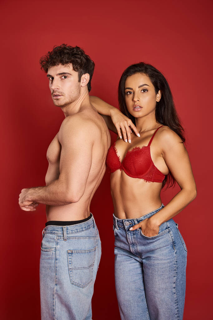 splendida giovane donna in reggiseno in posa con mano in tasca di jeans vicino a camicia fidanzato sul rosso - Foto, immagini