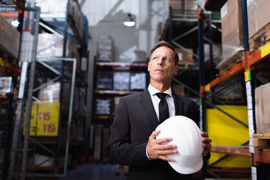 pensive middle aged businessman in suit holding hard hat in warehouse, professional headshot - Photo, Image