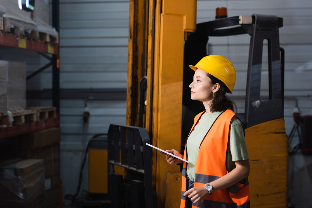 trabalhador armazém feminino em chapéu duro e colete de segurança segurando tablet digital perto de empilhadeira, carga - Foto, Imagem