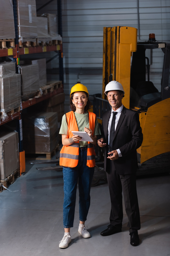 glücklicher Vorgesetzter in Anzug und Hut, der mit einer Mitarbeiterin auf dem Tablet steht, logistische Planung - Foto, Bild
