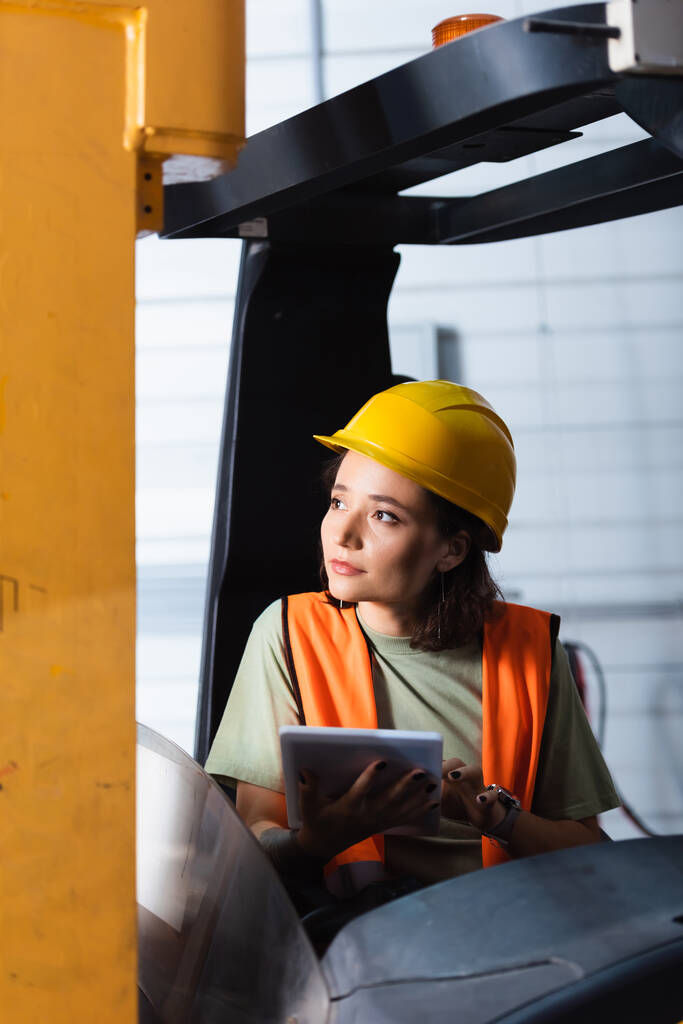 trabajadora de almacén pensativa en sombrero duro y chaleco de seguridad usando tableta y sentada en carretilla elevadora - Foto, imagen