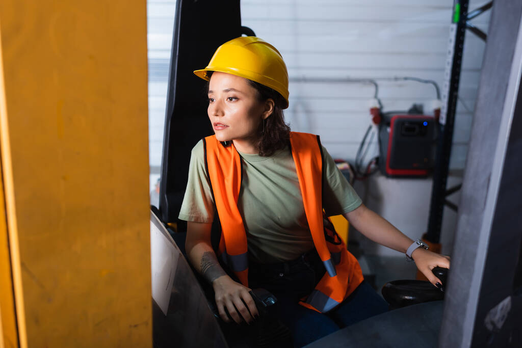 Sert şapka ve güvenlik yeleği giymiş düşünceli bir kadın forklift operatörü depoya bakıyor. - Fotoğraf, Görsel