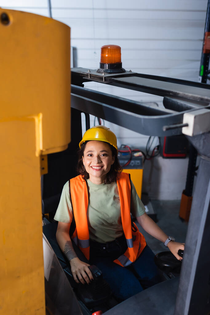 cheerful female forklift operator in hard hat and safety vest looking at camera in warehouse, cargo - Photo, Image