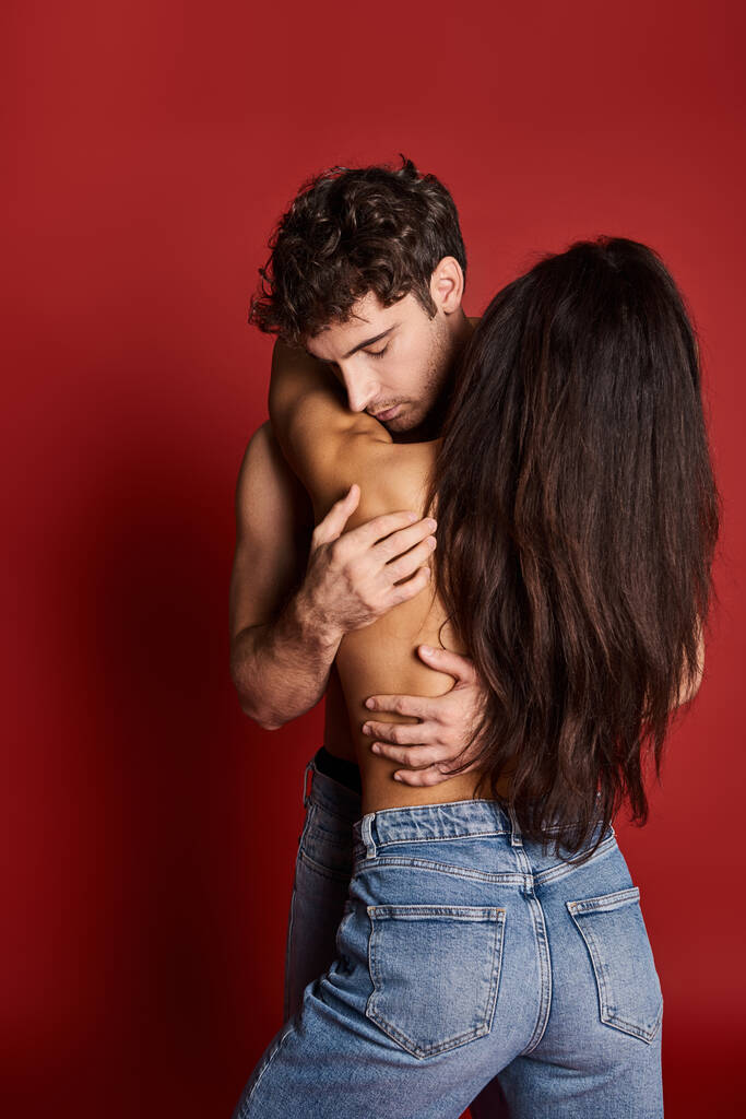 vista posterior de la mujer joven sin camisa con el pelo moreno abrazando hombre muscular en fondo rojo - Foto, imagen