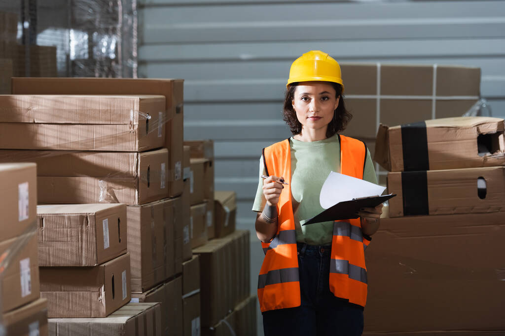 mujer trabajadora de almacén en casco duro sujetando portapapeles y mirando a la cámara, gestión logística - Foto, imagen