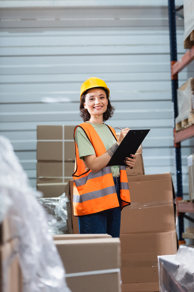cheerful female warehouse worker in hard hat and safety vest holding clipboard checking inventory - Photo, Image