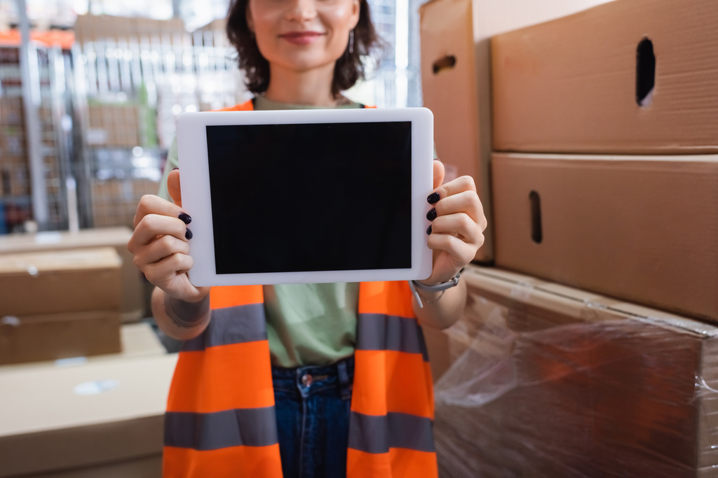 bijgesneden en glimlachende vrouwelijke magazijnmedewerker in veiligheidsvest met digitale tablet met blanco scherm - Foto, afbeelding