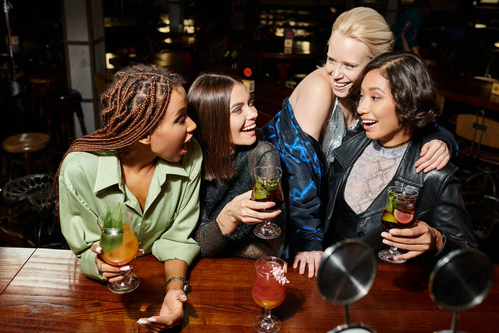 novias multiétnicas emocionadas y de moda con copas de cóctel hablando en el bar, ambiente vibrante - Foto, imagen