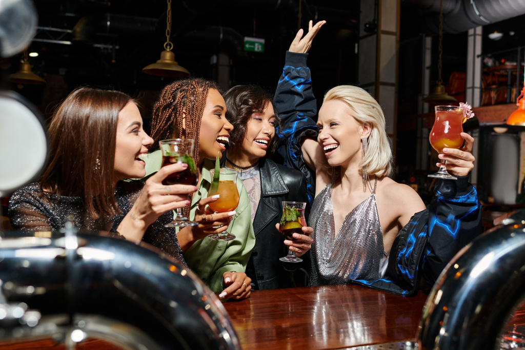 aufgeregte Frau prostet mit Cocktail und erhobener Hand bei Bar-Party mit multiethnischen Freundinnen zu - Foto, Bild