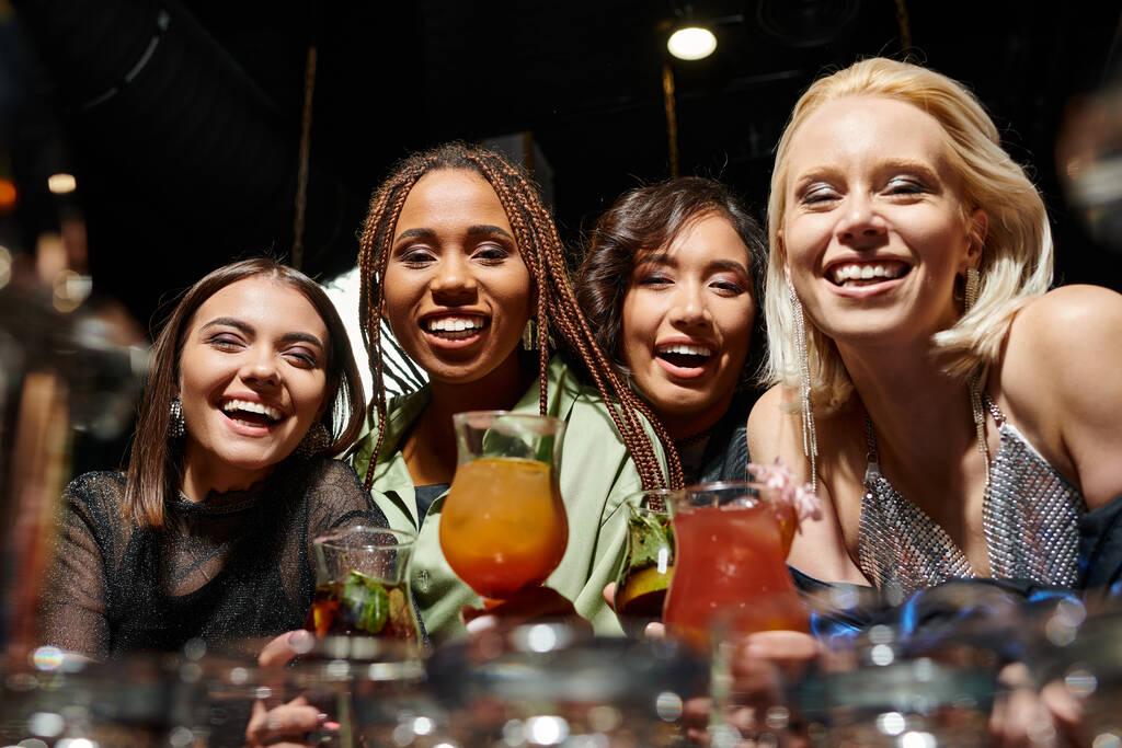 веселые многорасовые подружки, смотрящие в камеру рядом с вкусными коктейлями в баре, досуге и веселье - Фото, изображение