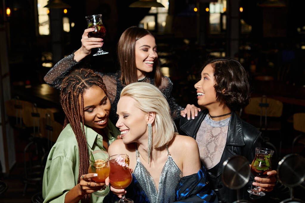 mulheres elegantes alegres que clinking copos de coquetel perto de amigas multiétnicas durante a festa no bar - Foto, Imagem