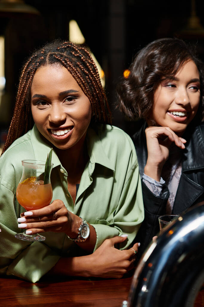 mulher americana africana elegante com coquetel de vidro olhando para a câmera perto de namorada asiática no bar - Foto, Imagem