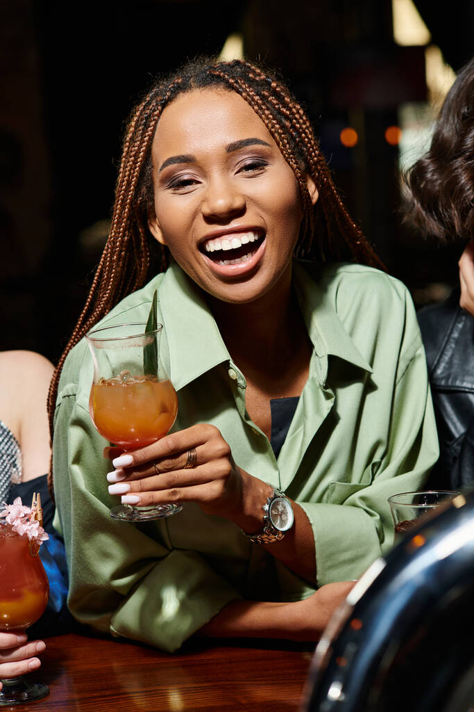 Kokteyl bardağıyla bardaki kız arkadaşlarının yanında gülen heyecanlı ve modaya uygun Afrikalı Amerikalı kadın. - Fotoğraf, Görsel