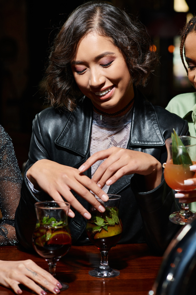 молода і стильна азіатка посміхається біля коктейльного скла і мультикультурних подружок в барі - Фото, зображення