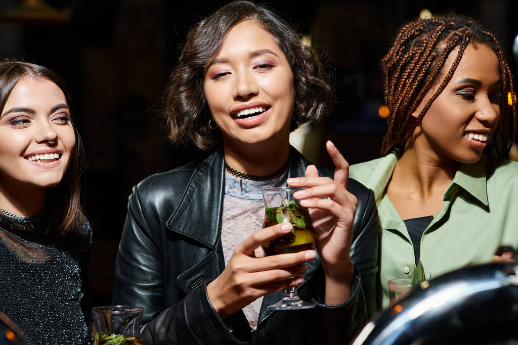 mladá a trendy asijské žena s koktejlovými brýlemi s úsměvem v blízkosti multietnické přítelkyně v nočním baru - Fotografie, Obrázek