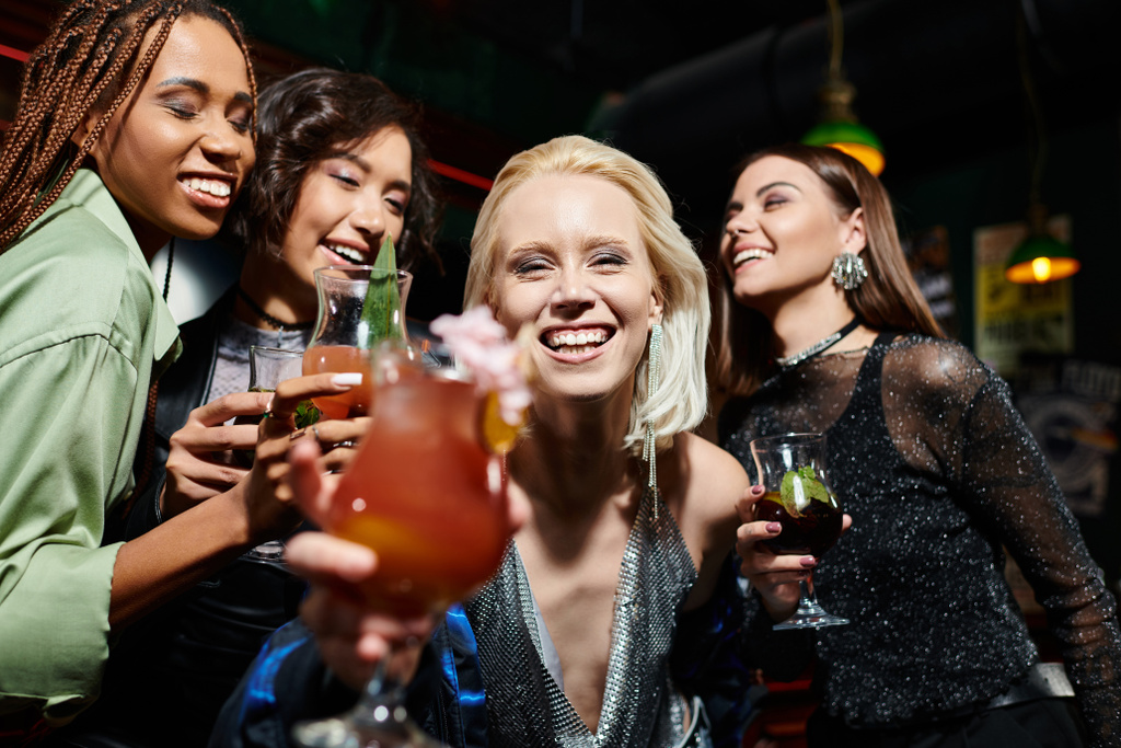 happy blonde woman with glass of cocktail smiling at camera near multiethnic girlfriends in bar - Photo, Image