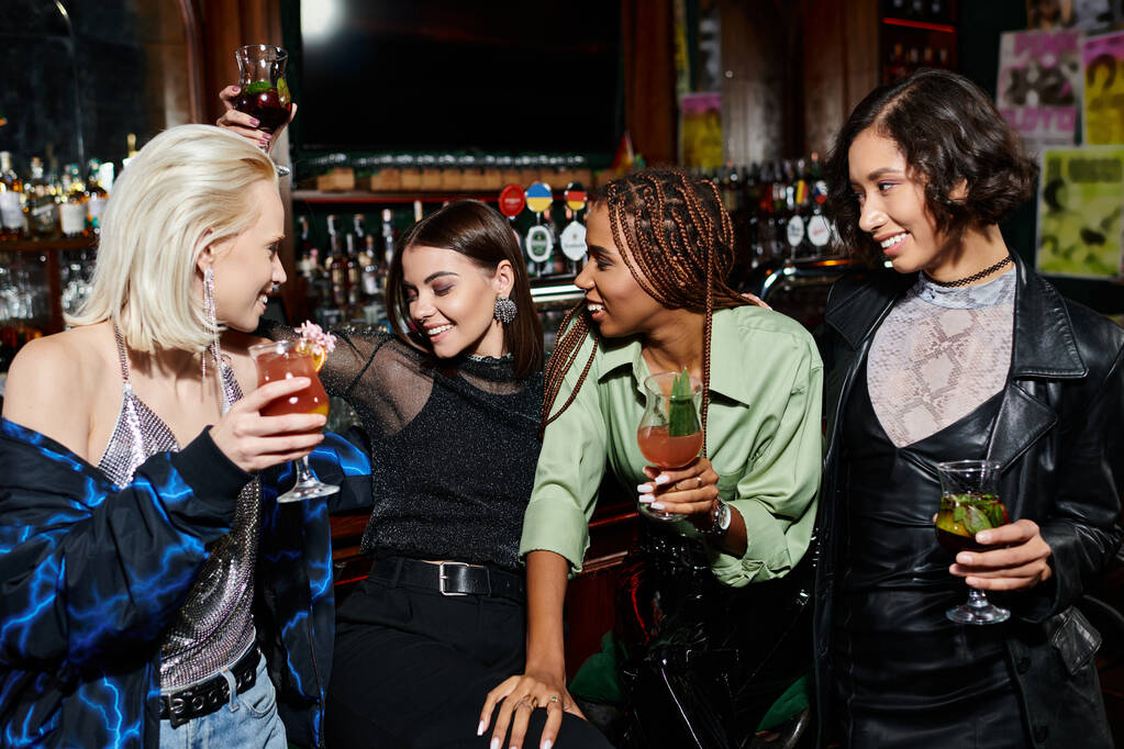 vidám, többnemzetiségű női barátok koktélokat tartanak beszélgetés közben a modern éjszakai bárban - Fotó, kép