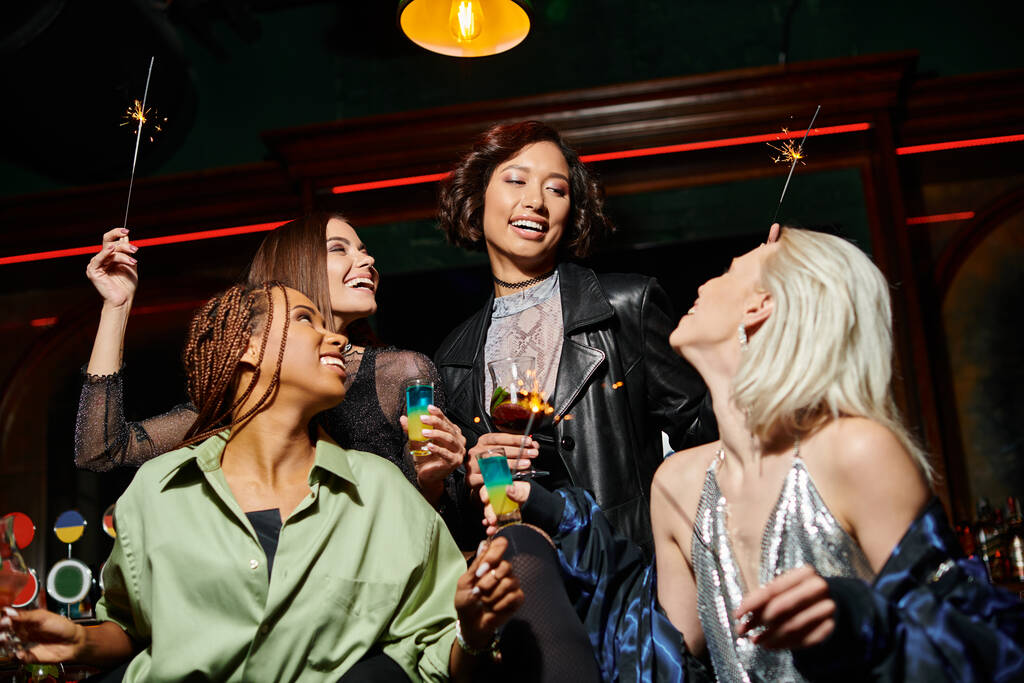 Улыбающиеся и стильные многорасовые подружки с коктейлями и блестящими искорками в баре, праздничная вечеринка - Фото, изображение