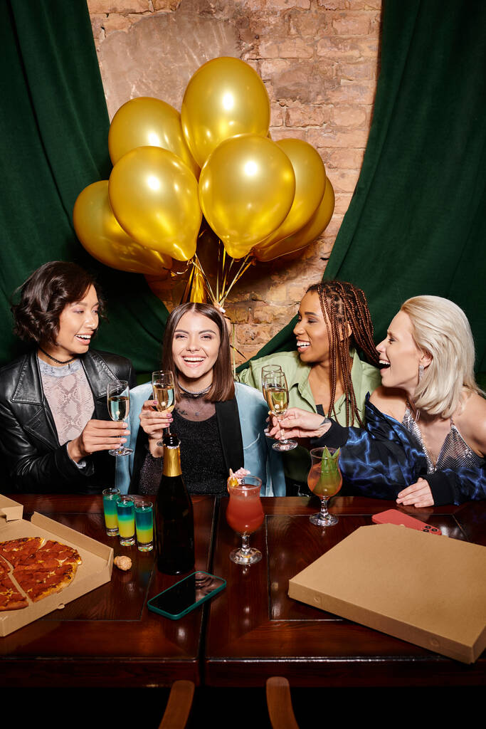 cheerful multiracial fashionistas with champagne near cocktails and golden balloons in bar, birthday - Photo, Image