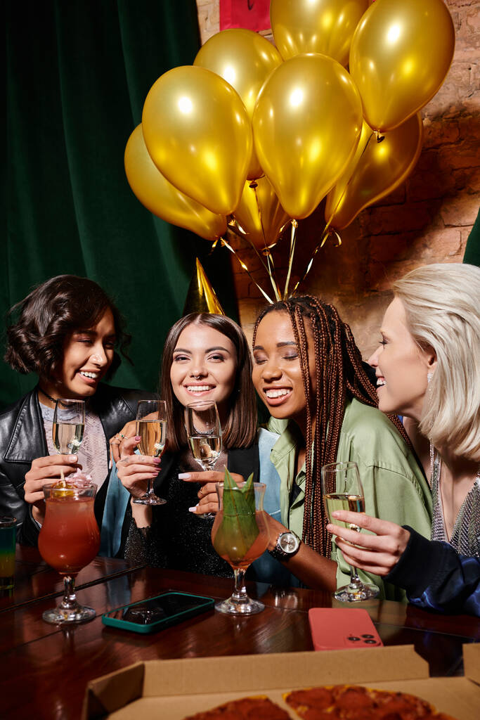 счастливая женщина в шляпе для вечеринок с шампанским рядом с многонациональными подругами и воздушными шариками на день рождения в баре - Фото, изображение