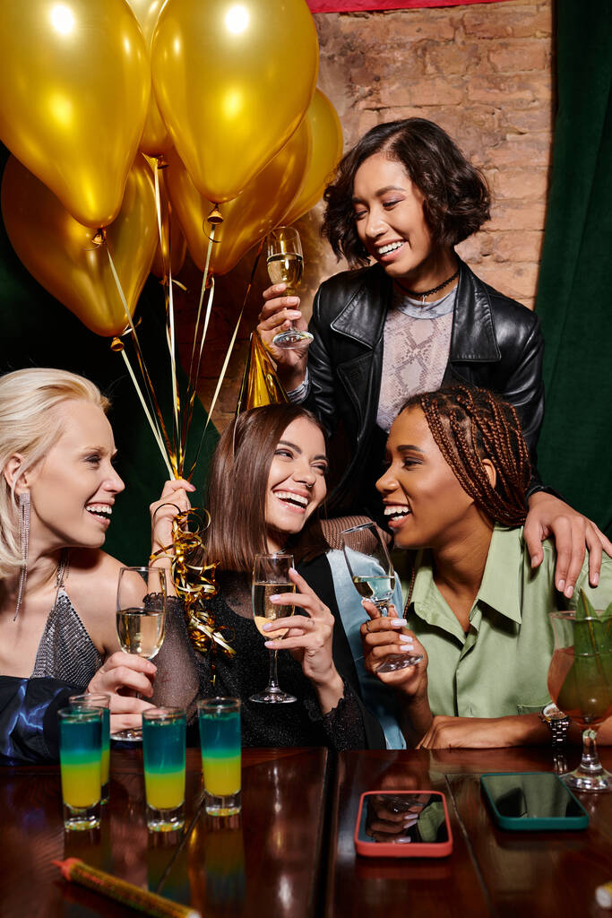novias multiétnicas de moda con champán sonriendo cerca de cócteles y globos de oro, cumpleaños - Foto, Imagen