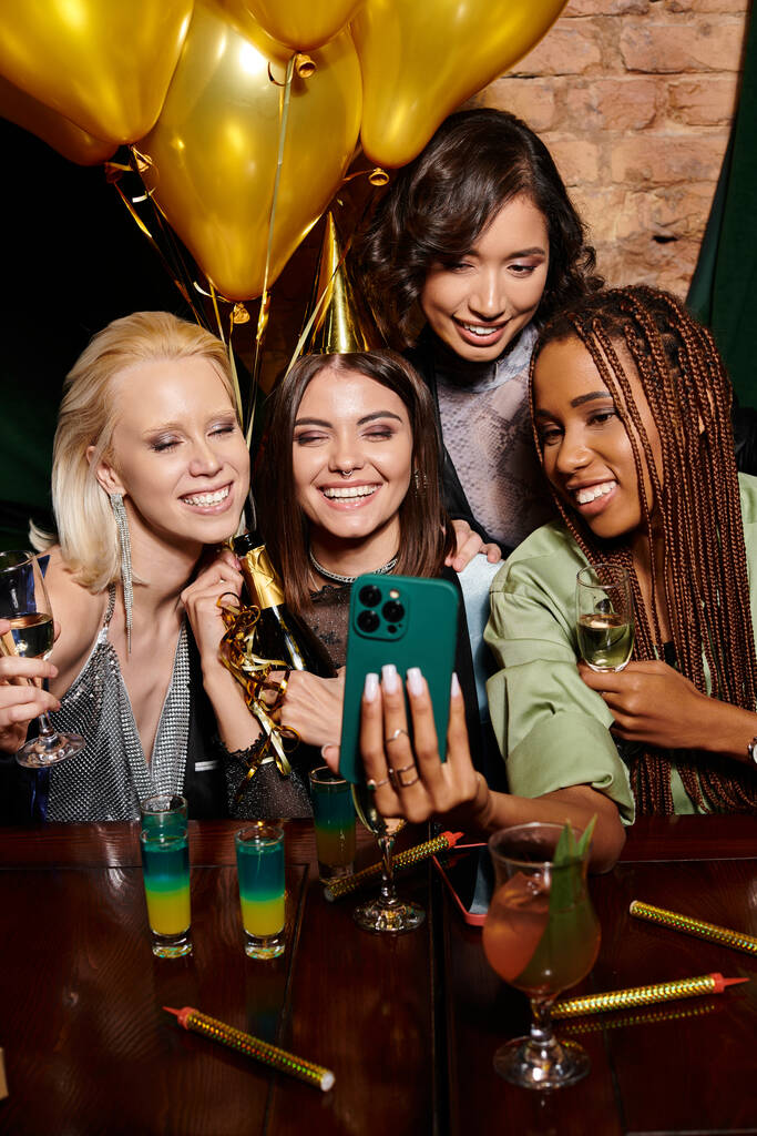 femme heureuse dans le chapeau de fête prendre selfie avec des copines multiraciales élégantes, fête d'anniversaire dans le bar - Photo, image