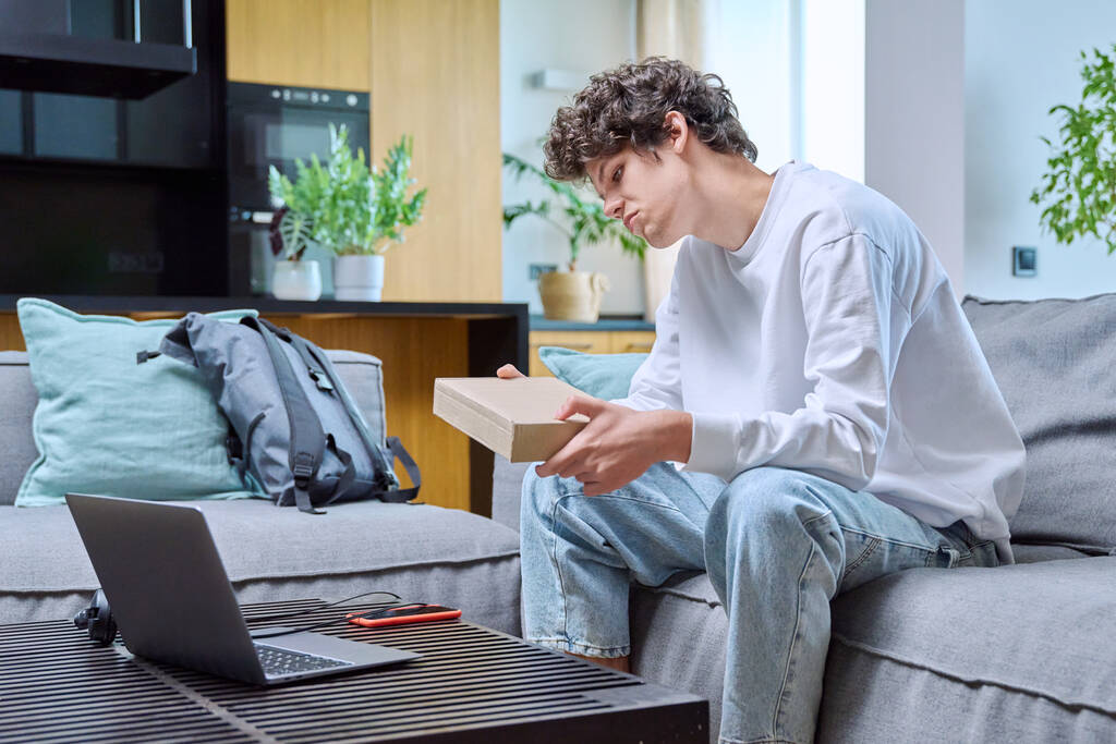 Jeune homme déballer un achat en ligne, assis sur le canapé à la maison. Achats en ligne sur Internet, livraison, concept jeunesse - Photo, image