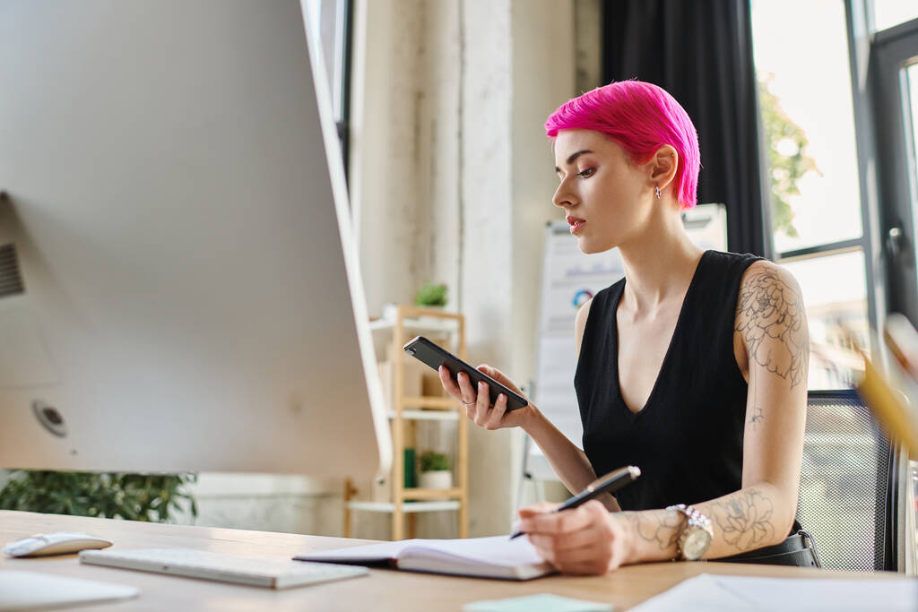 attractive pensive businesswoman with pink hair and tattoos looking at phone while taking notes - Photo, Image