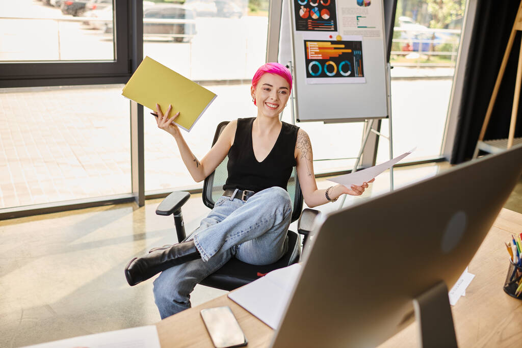 веселая молодая работница улыбается компьютерной камере, держа в руках свои заметки, бизнес-концепцию - Фото, изображение