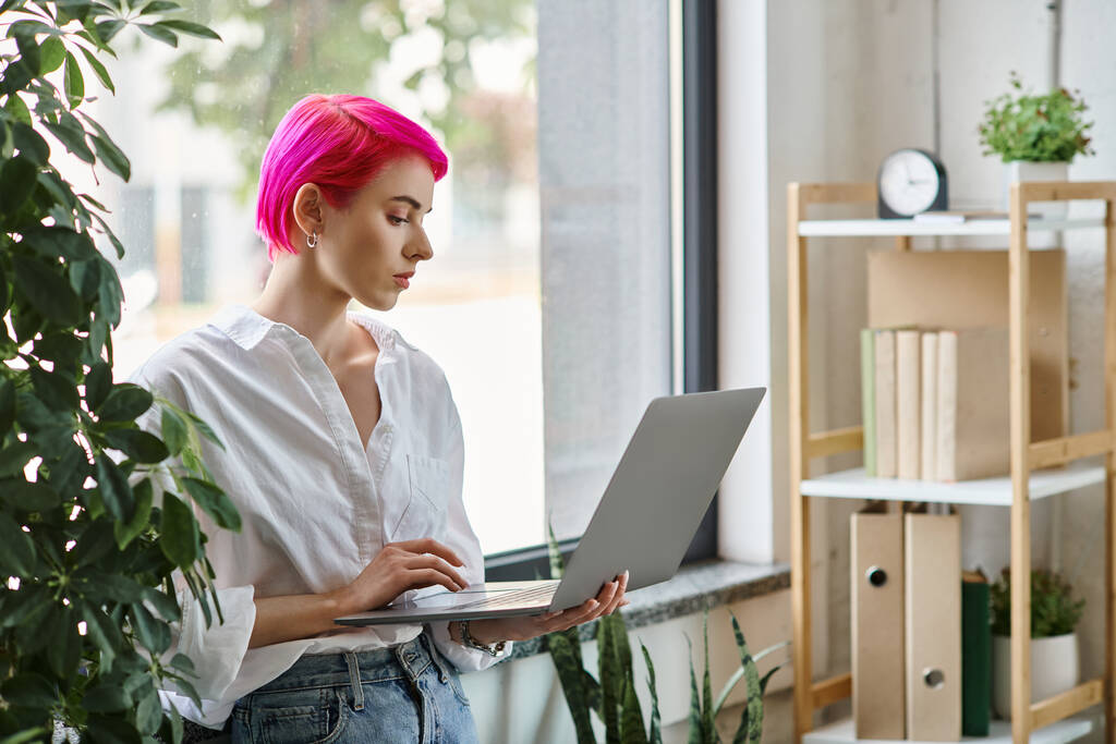 красива рожева волохата бізнес-леді в білій сорочці і джинсах, що працюють на її ноутбуці, стоячи - Фото, зображення