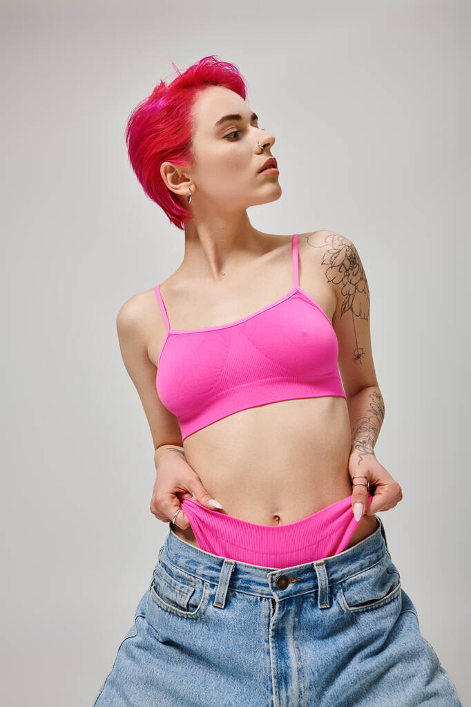татуйована стильна жінка з рожевим волоссям позує у верхній частині культури і витягує трусики з джинсів на сірий - Фото, зображення