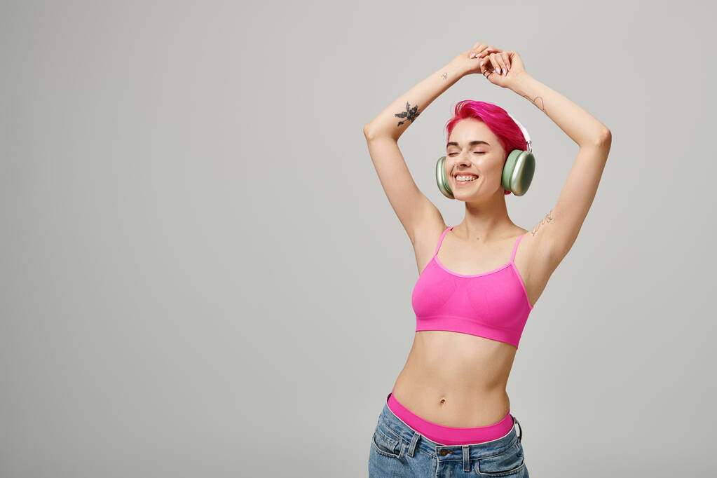 mujer feliz con pelo rosa escuchando música en auriculares inalámbricos sobre fondo gris, manos levantadas - Foto, Imagen