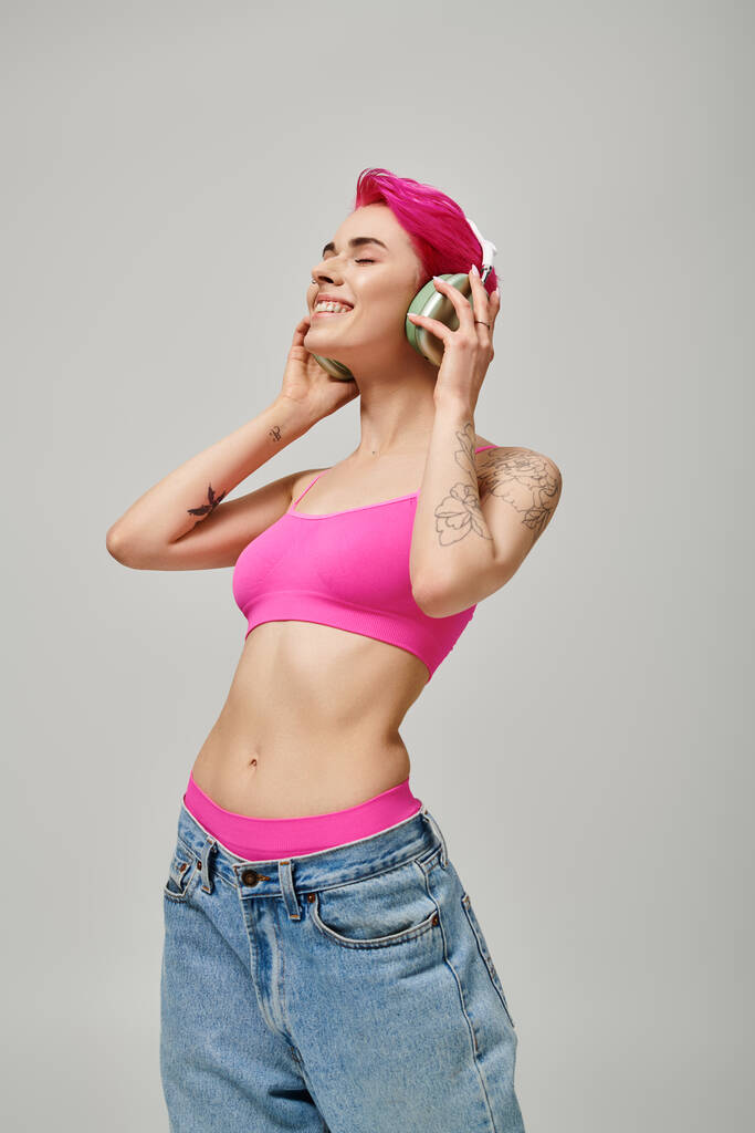 femme élégante tatouée avec des cheveux roses écoutant de la musique dans des écouteurs sans fil sur fond gris - Photo, image