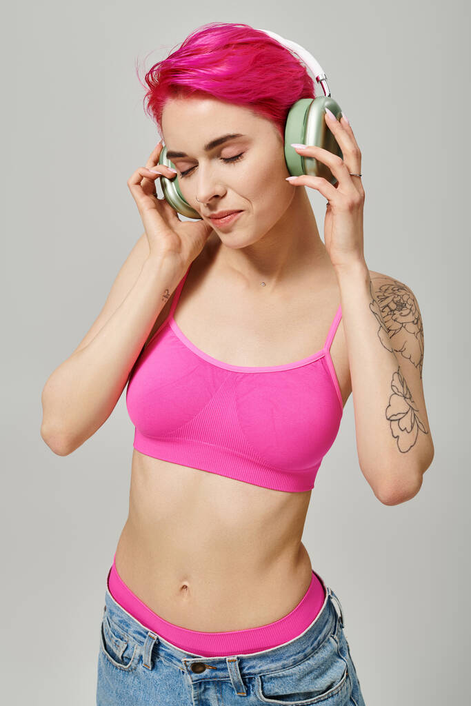 tatuoitu nuori nainen vaaleanpunaiset hiukset kuuntelevat musiikkia langattomissa kuulokkeissa harmaalla taustalla - Valokuva, kuva