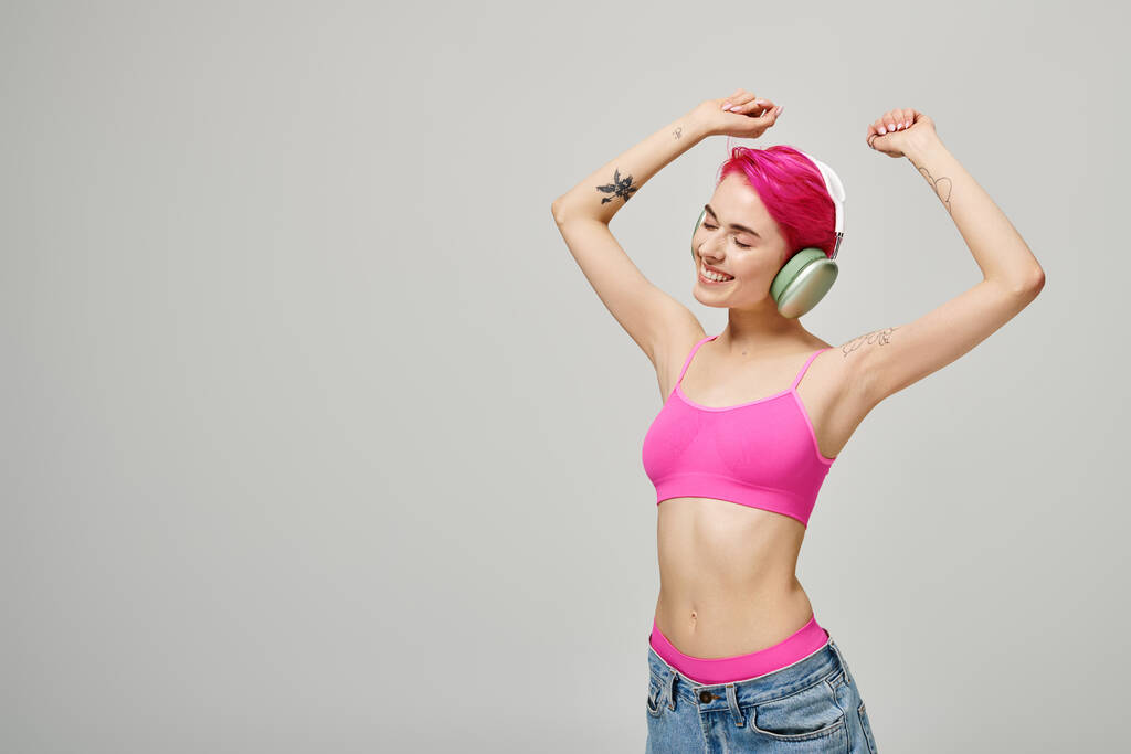 веселая и пронзительная женщина с розовыми волосами слушает музыку в беспроводных наушниках на сером фоне - Фото, изображение