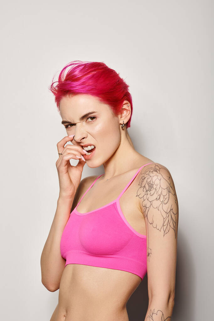 érzelmi tetovált fiatal nő rózsaszín haj pózol a termés felső és harapás szög szürke háttér - Fotó, kép