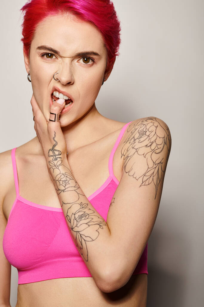tattooed and pierced woman with pink hair and posing in crop top biting finger on grey background - Photo, Image