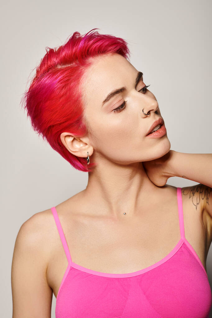 портрет татуированной и пронзительной молодой женщины с розовыми волосами, смотрящей в сторону на сером фоне - Фото, изображение